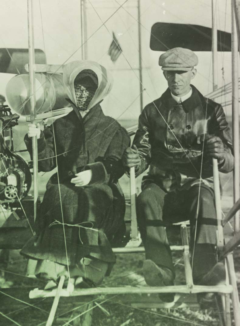 Man in leather jacket and cap sits beside woman in hat, veil, and long coat among the structure of an apparatus