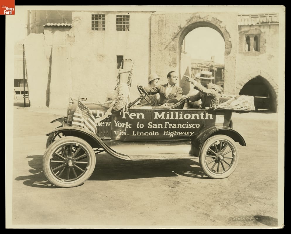 Mary Pickford and Douglas Fairbanks in the Ten-Millionth Ford Model T, 1924 Black-and-white photo of three people sitting in an early open car with text on side, parked in front of buildings and a masonry arch