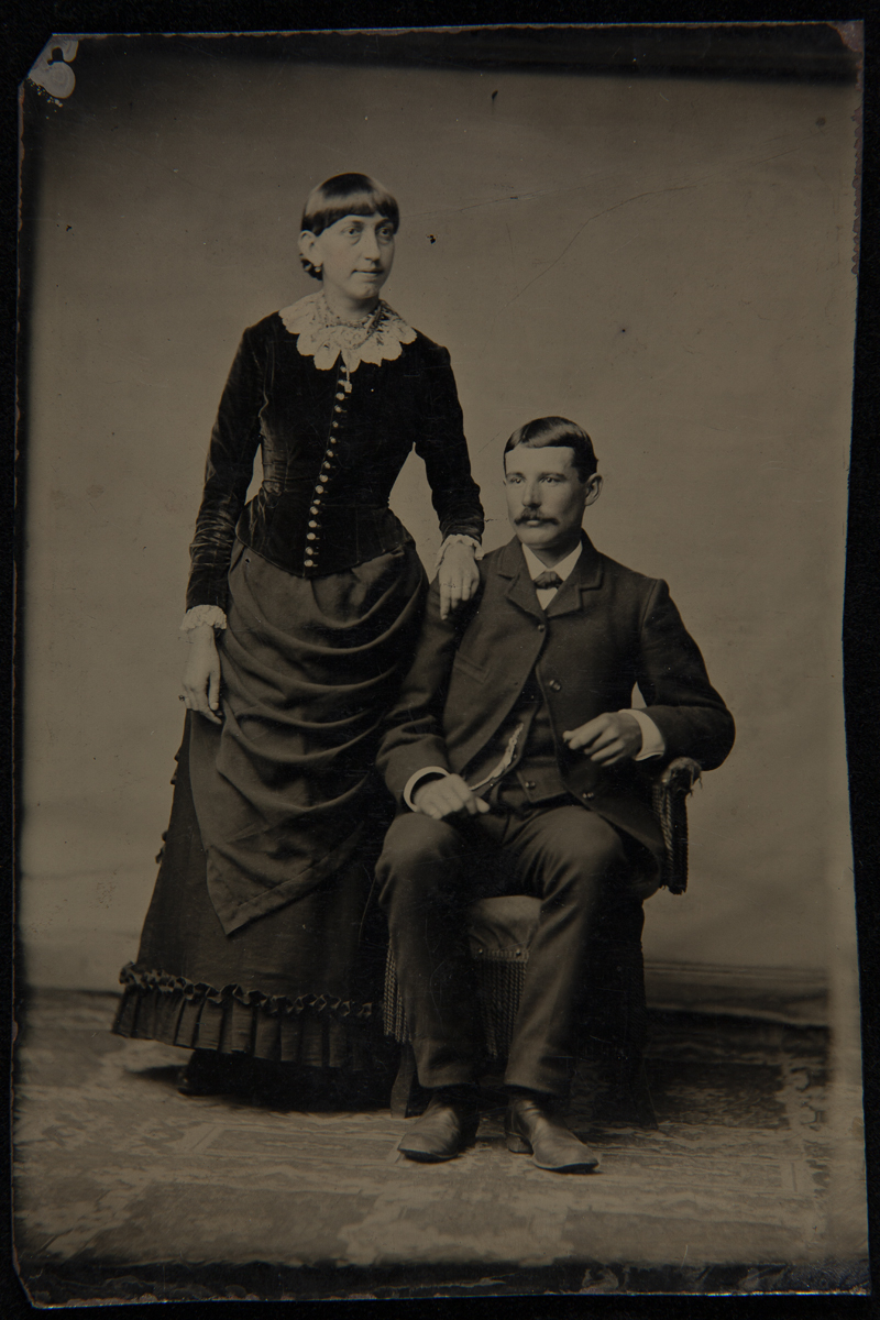 Woman with bangs wearing elaborate ensemble, standing with hand on shoulder of seated man wearing a suit