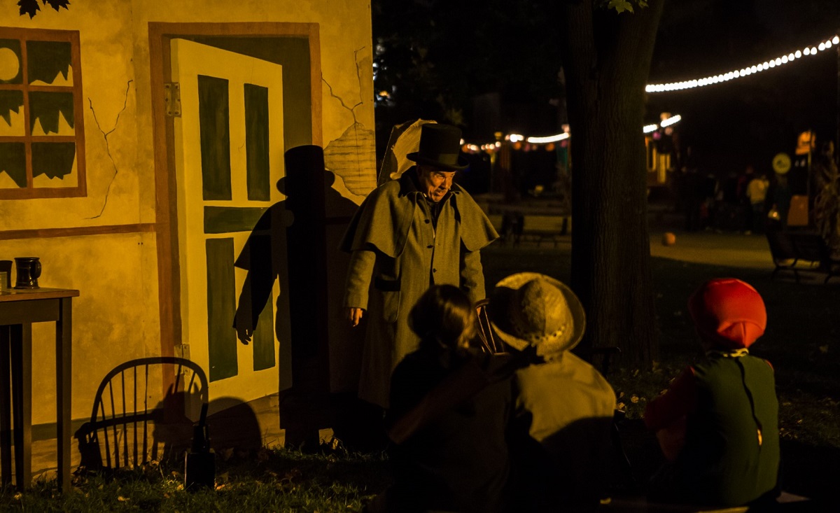 Man in overcoat and top hat stands in front of a backdrop of a building as seated people look on