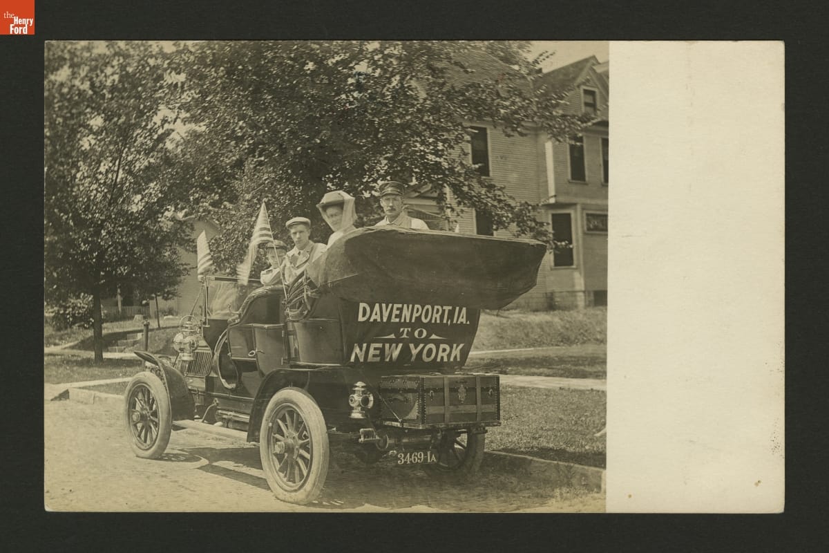 People sitting in an old-fashioned open car, with "Davenport, IA. to New York" written behind the rumble seat
