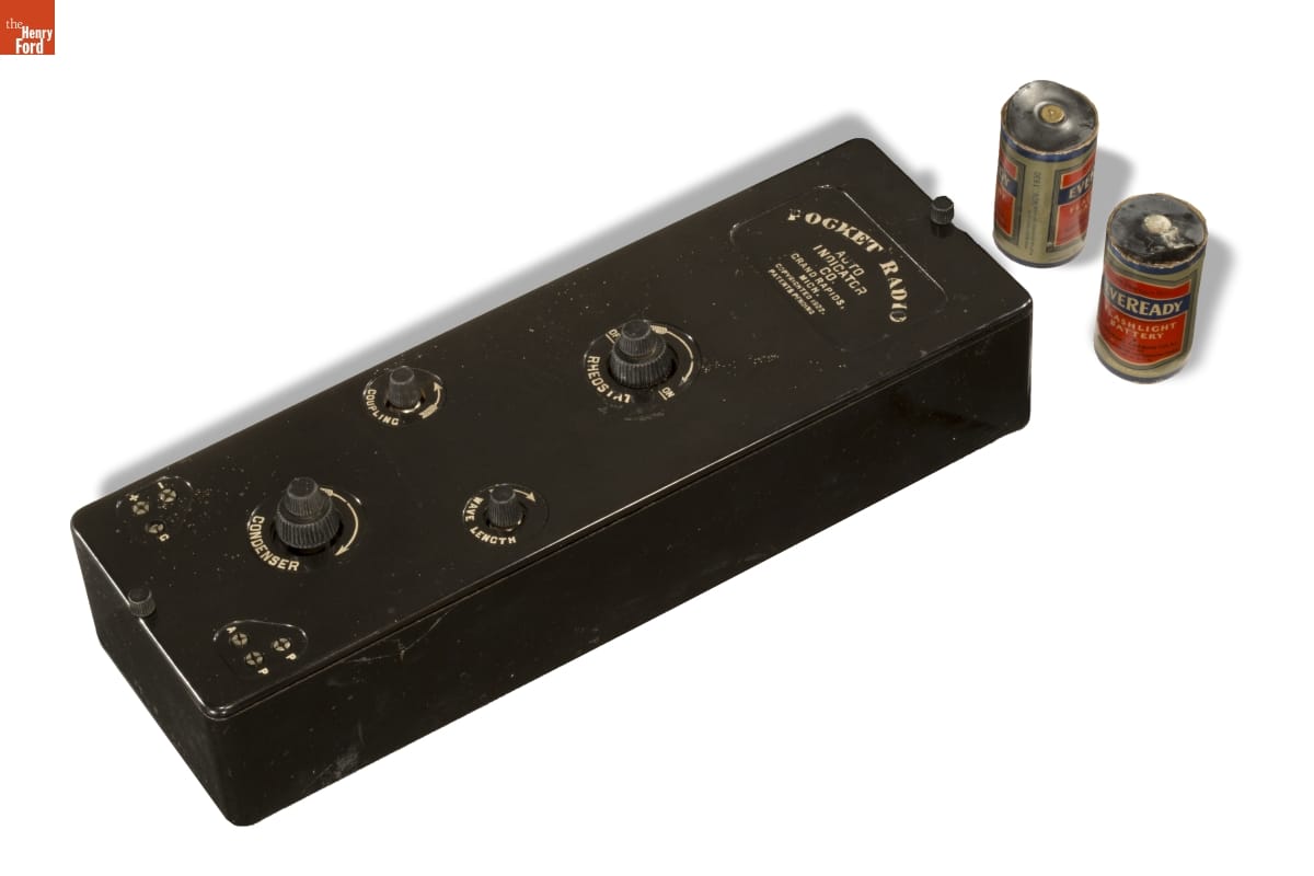 Black rectangle with text and several dials and knobs; two batteries next to it