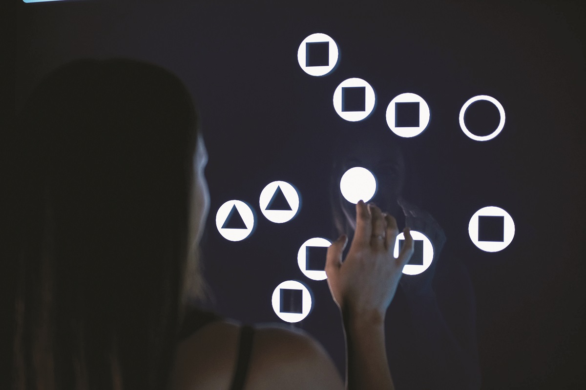 Woman stands with hand up to a black digital board with lighted circles, triangles, and squares within white circles