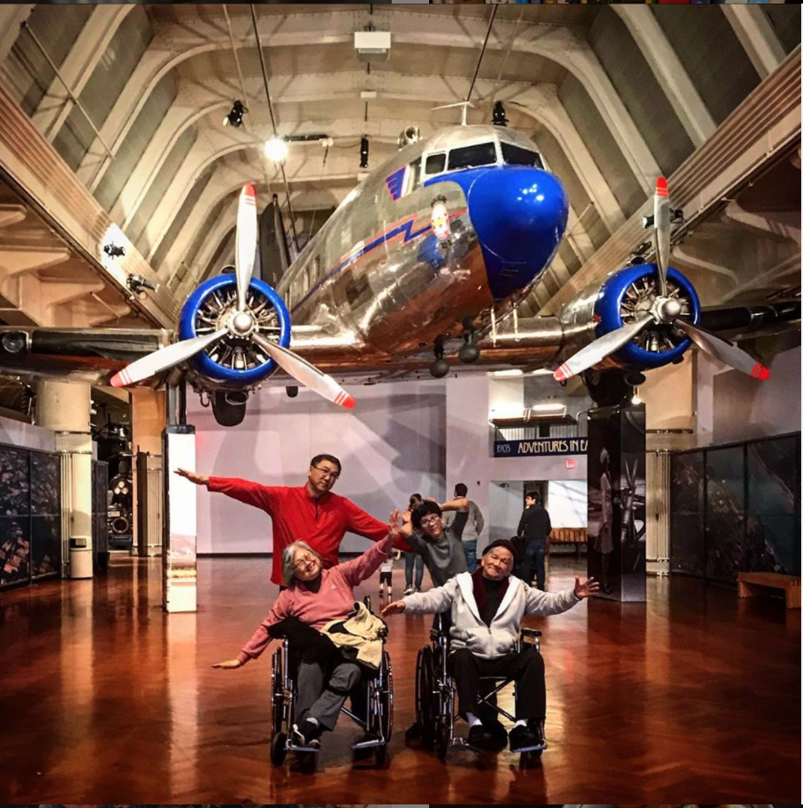 Visitors pose underneath the DC-3 in Henry Ford Museum of American Innovation. Four people (two in wheelchairs) pose with arms outstretched underneath a silver-and-blue airplane