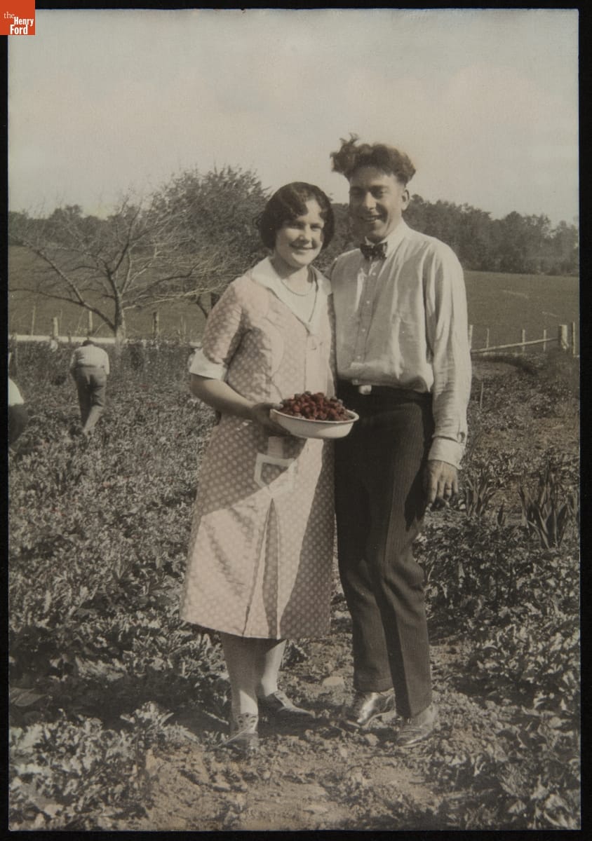 Partially colorized black-and-white photo of a woman and man standing next to each other in a field; the woman holds a bowl of strawberries