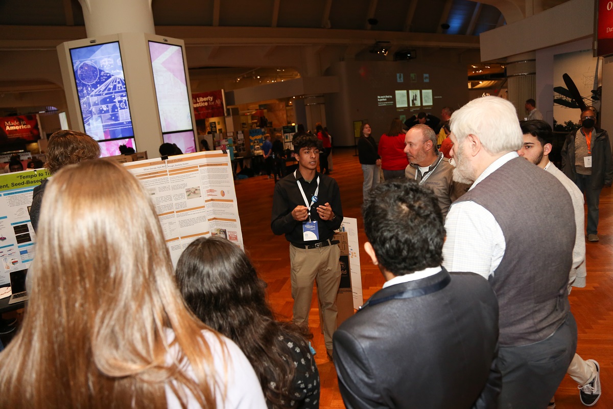 A young man stands by a trifold display board, talking to a group of people circled around him