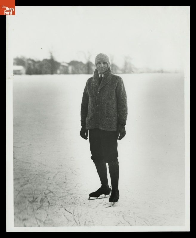 Man in cardigan with collar turned up, hat, knickers, and ice skates, on ice with trees and buildings in background