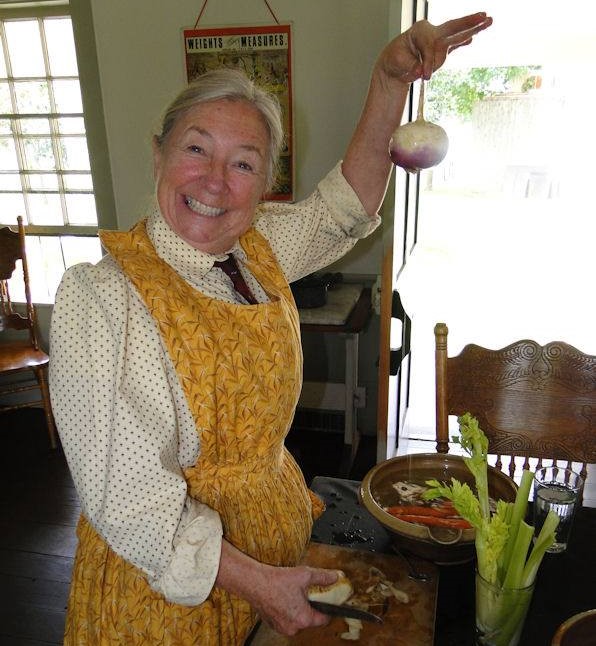 A smiling woman wearing an apron works in a kitchen, with a knife on a chopping board in one hand, holding up a turnip with the other hand