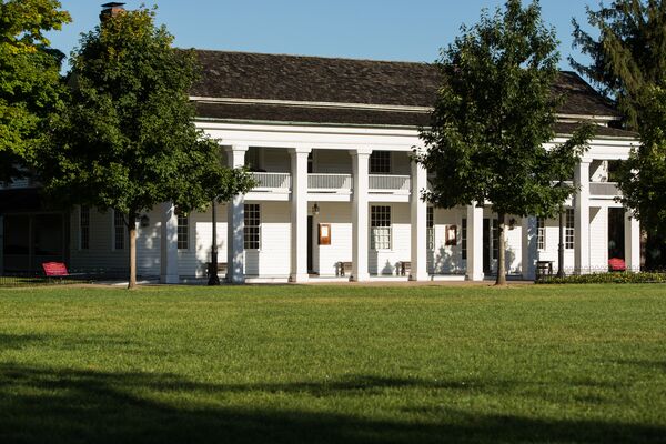 Two-story white building with columns and second-floor balcony with two small trees in front on a grassy lawn