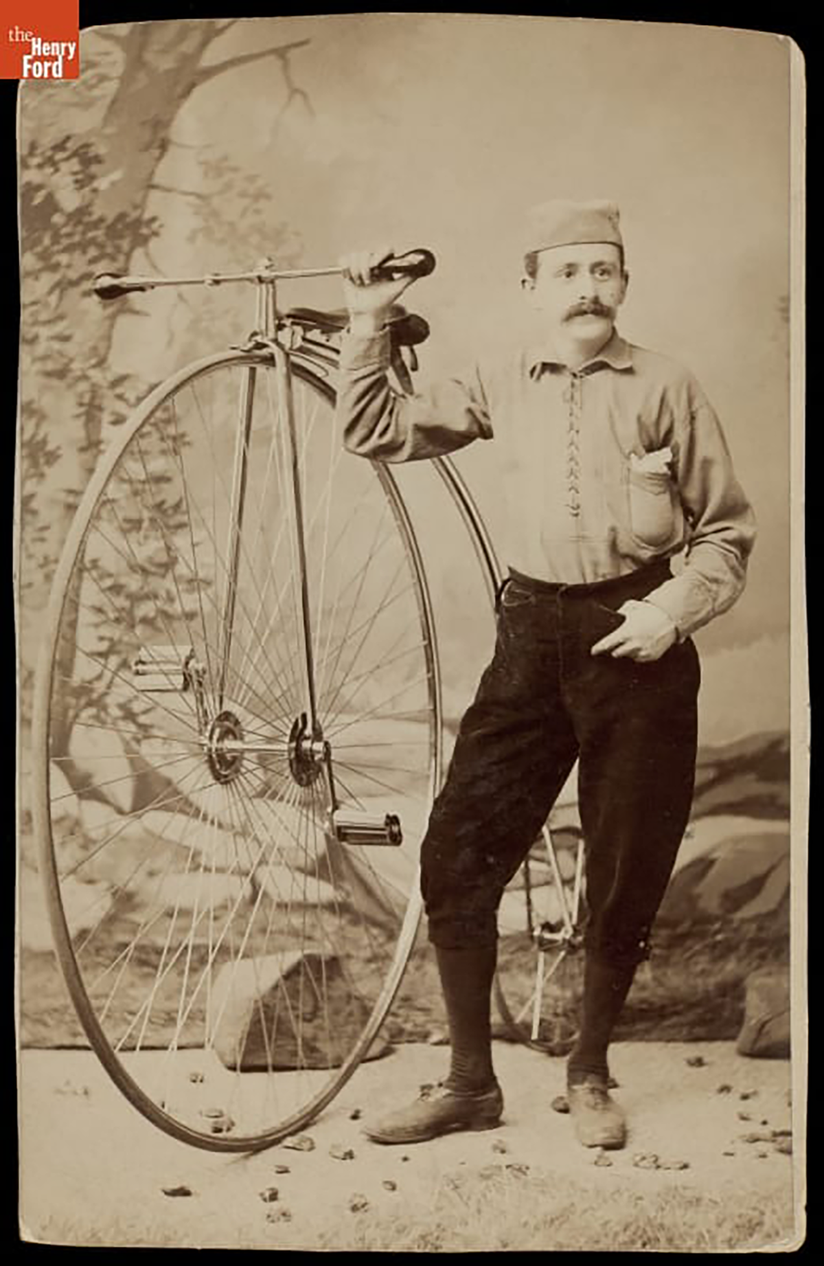 A man with a mustache standing next to a bicycle with a front wheel as tall as him