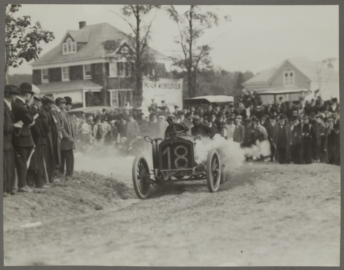 Early open race car careens around a corner on a dirt road, kicking up clouds of smoke with spectators looking on and houses in the background