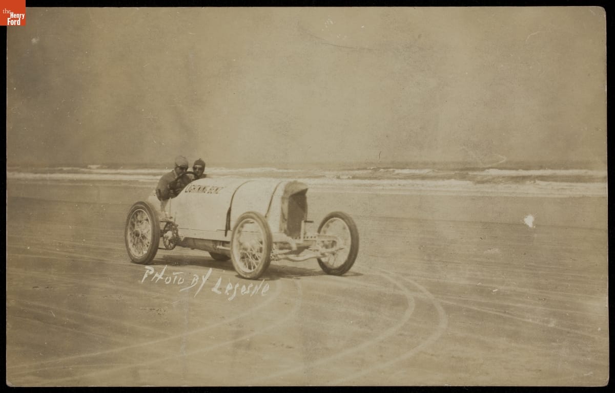 Sepia-tone photo of two men in early open race car on beach with ocean behind them; also contains text