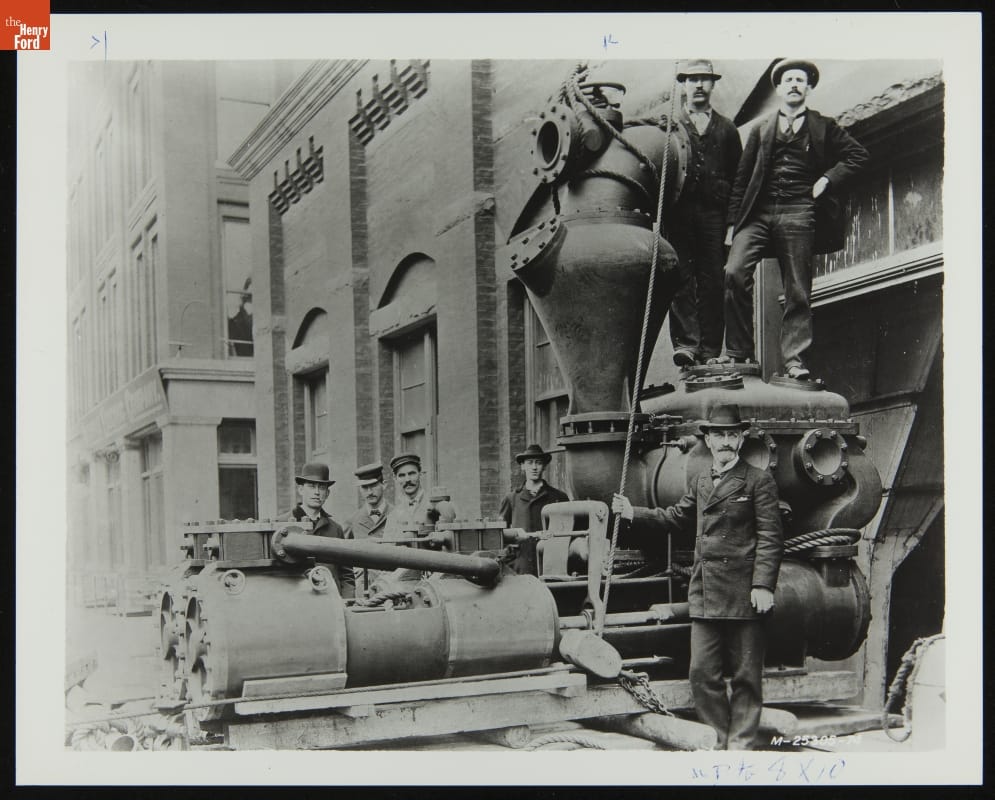 Black-and-white photograph of men standing among machinery outside a large brick building